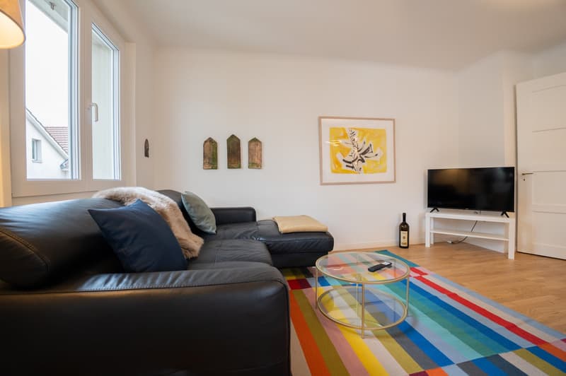 Spacious living room with large windows, black leather sofa, colorful area rug, glass coffee table, and wall decor including framed artwork and wooden shelves.
