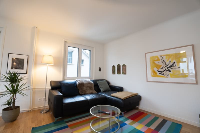 Spacious living room with white walls, hardwood floors, large windows, black leather sofa, glass coffee table, colorful area rug, potted plant, and framed artwork on the walls.