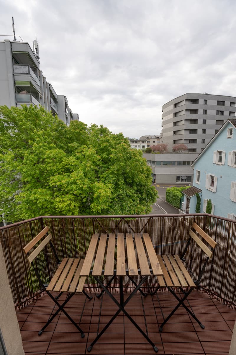 Wooden deck with a table and chairs, surrounded by a bamboo fence. The balcony overlooks a residential area with apartment buildings and trees.