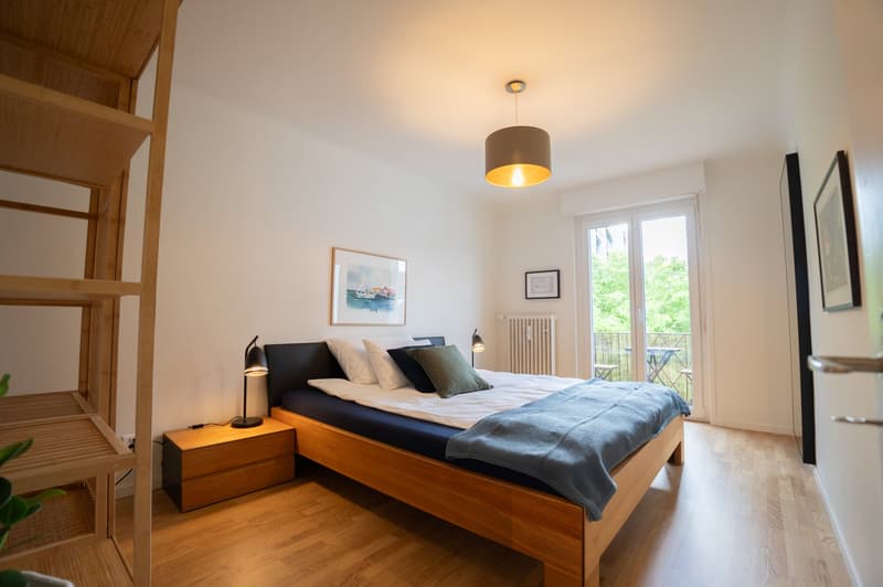 Spacious bedroom with wooden floor, white walls, large windows leading to a balcony, a wooden bed frame with a gray bedspread, nightstands, and a pendant light fixture above the bed.