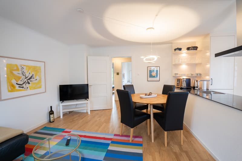 The image shows an open-plan living room and kitchen area. The living room has a large TV mounted on the wall, and a colorful patterned rug on the hardwood floor. The dining area features a round wooden table with black leather chairs. The kitchen has mod