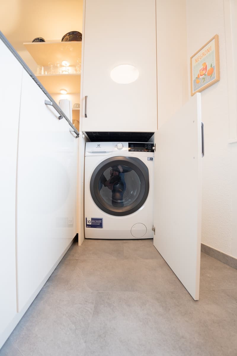 The image shows a small laundry room with a washing machine and shelves above it. The room has white walls and a tiled floor.
