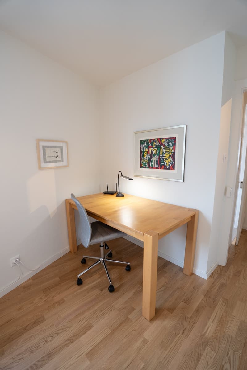 The image shows a simple and minimalist home office setup. The room has white walls and a wooden floor. There is a wooden desk with a chair in front of it. On the desk, there is a framed artwork on the wall above it.