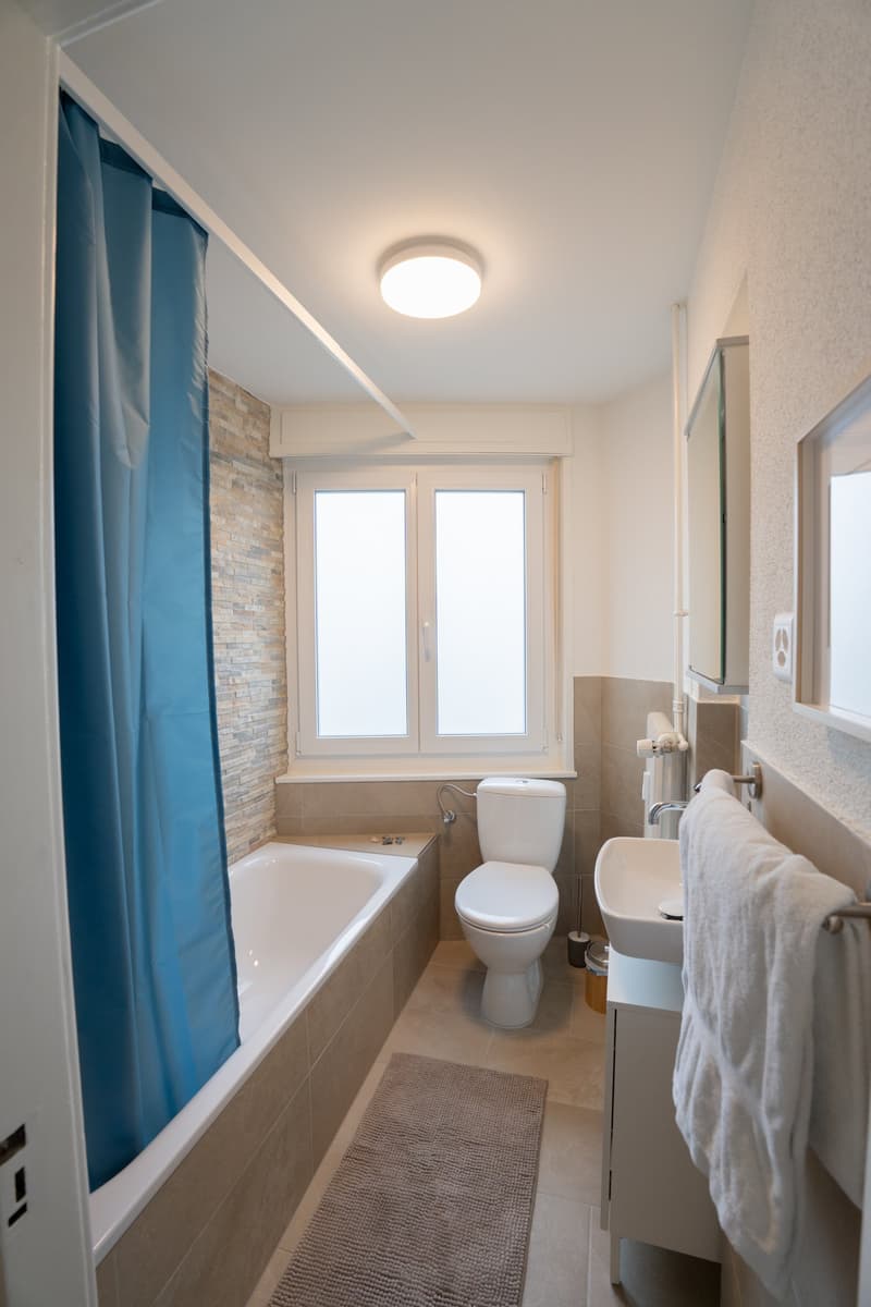 The image shows a modern bathroom with a bathtub, toilet, vanity, and large windows. The bathroom has a clean, minimalist design with white walls, beige tiles, and a blue shower curtain. There is a towel rack and a small rug on the floor.