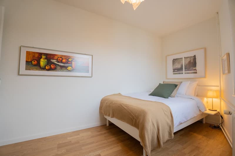 The image shows a cozy bedroom with a white wall, hardwood floors, a large framed painting on the wall, and a bed with a beige blanket and green accent pillow. There is also a lamp on a side table and some additional artwork on the wall.