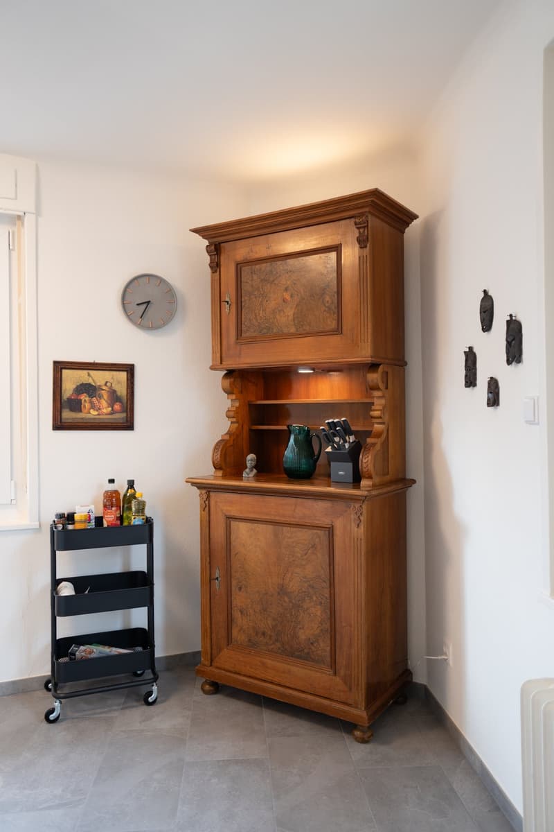The image shows a kitchen or dining area with a large, ornate wooden cabinet or hutch as the central piece of furniture. The cabinet has a decorative top and shelves for displaying items. There is also a wall clock, a framed painting, and a black metal ut