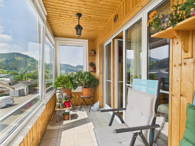 Spacious balcony with wooden ceiling, large windows offering scenic mountain views, potted plants, and comfortable outdoor furniture