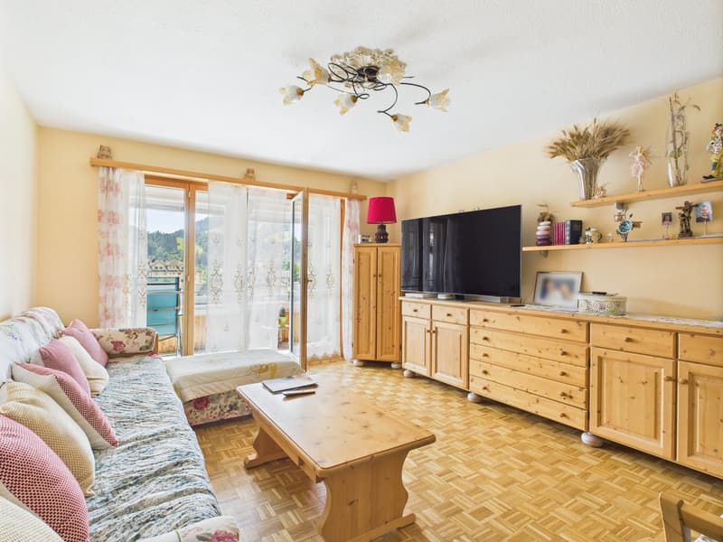 Spacious living room with large windows, wooden furniture including a TV cabinet and coffee table, floral patterned sofa, and a decorative ceiling light fixture