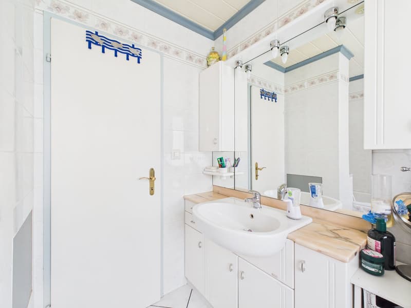 Bathroom with white walls, tiled floor, vanity sink, mirror, and shelving. There is a private washing machine visible.