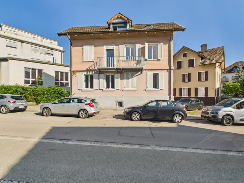 A multi-story residential building with multiple cars parked in front, white shutters, brown roof, balcony on the second floor.