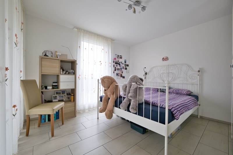 Bedroom with white bed, chair, cabinet, stuffed animals, picture on wall, window with curtain