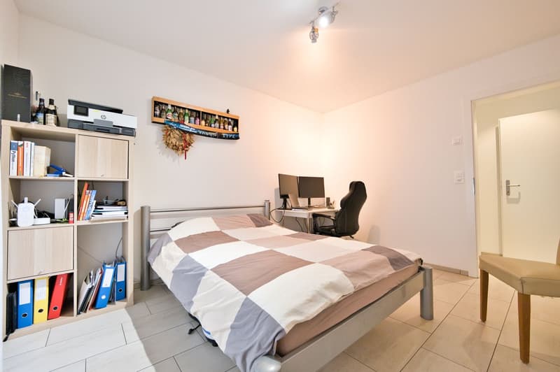 A bedroom with a bed, a desk with a computer, a chair, a shelf with books, a printer, wine bottles, and a wreath on the wall.