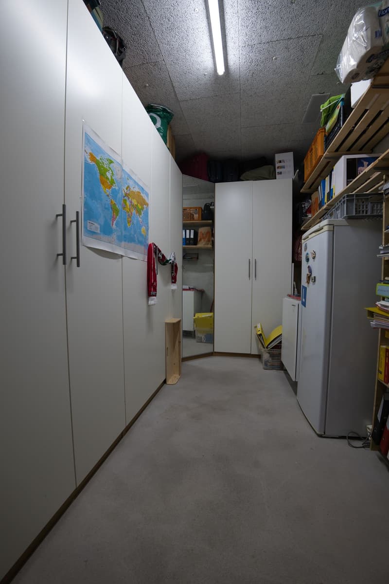 Storage room with wooden cabinet, a white fridge, map on the wall and some shelves, a mirror and white tiles floor.
