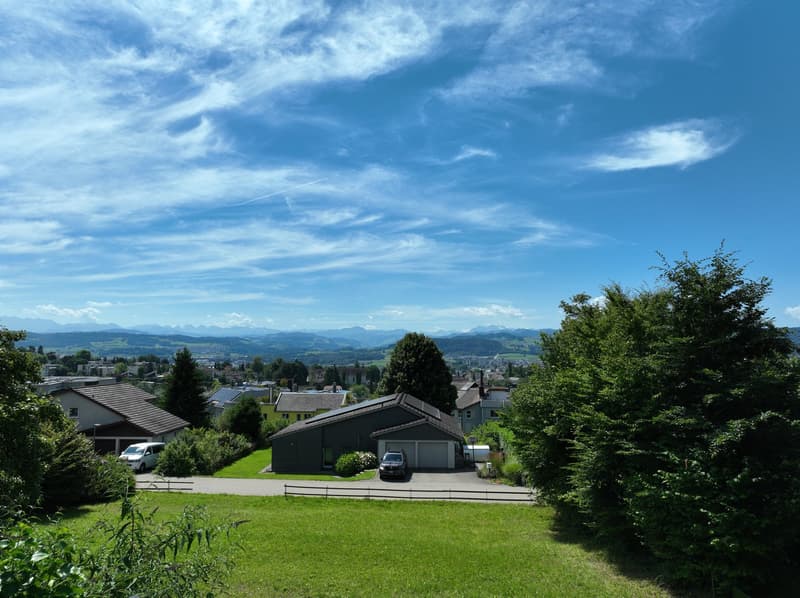 The image shows a residential area with houses, trees, and a grassy lawn in the foreground. The houses have dark roofs and there are cars parked in the area. The background features a scenic view of mountains and a blue sky with wispy clouds.
