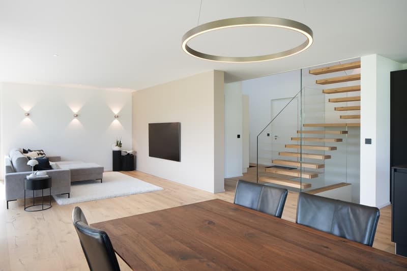 modern living room with wooden floor, white walls, gray sofa, dining table, glass staircase, round ceiling lamp