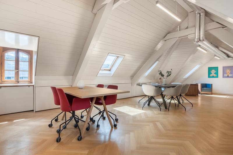 Wooden herringbone floors, two desks, red chairs, wooden table, white ceiling beams, skylights, windows