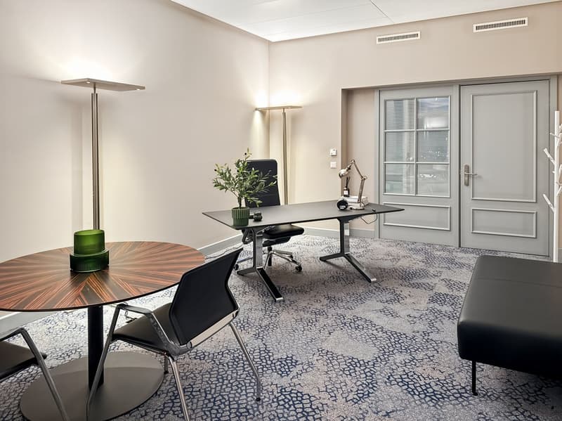 Modern office space with large windows, adjustable desk, comfortable chairs, and decorative elements like plants and lamps.