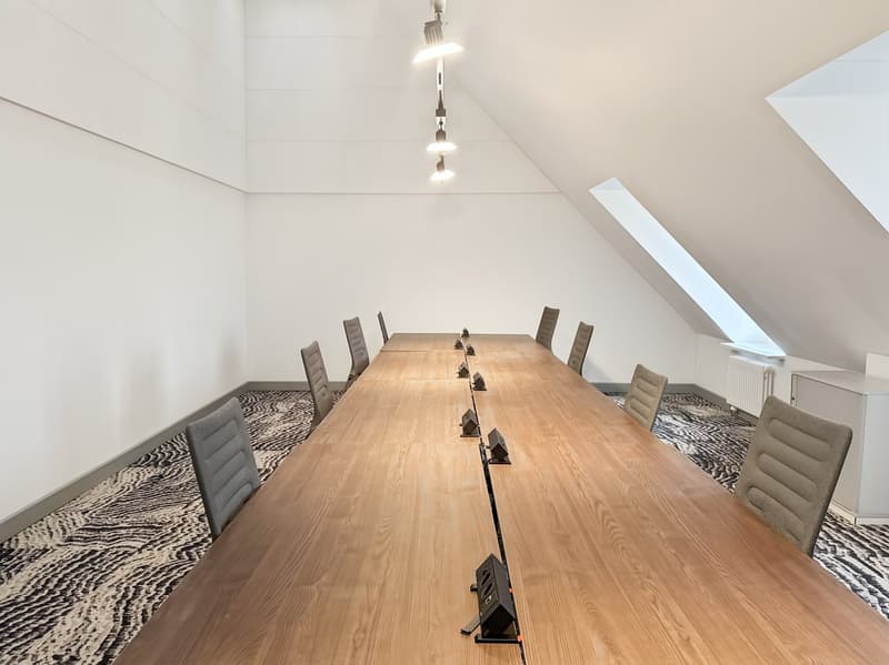 Long wooden conference table, grey chairs, tiled flooring, angled ceiling with lamps