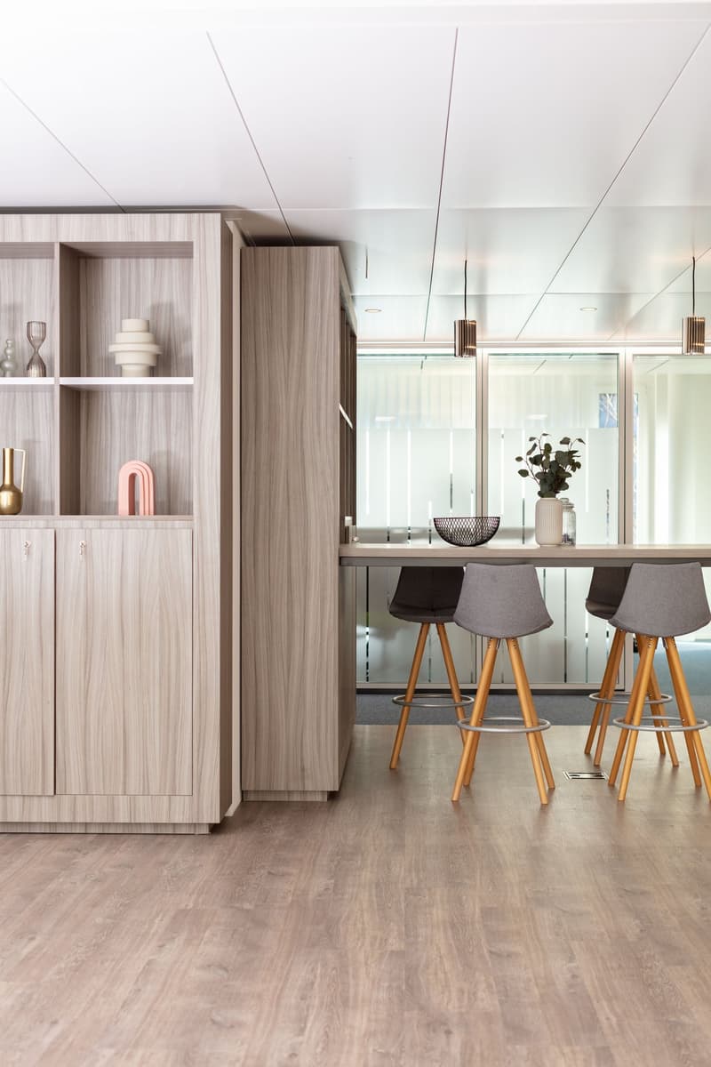 Large, open area with bar counter, wooden flooring, modern furniture, high ceiling with lights, glass windows, plants, shelves with decorations, and decorative items.