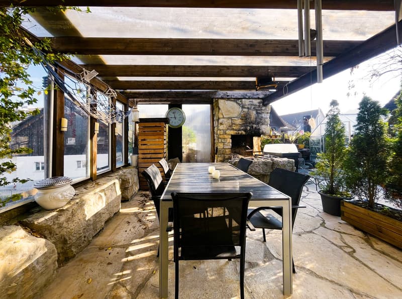 Outdoor dining area with a wooden table and chairs, stone bench, and a stone wall. There is a fireplace, a clock, and a lamp on the wall. Potted plants and trees are also present.