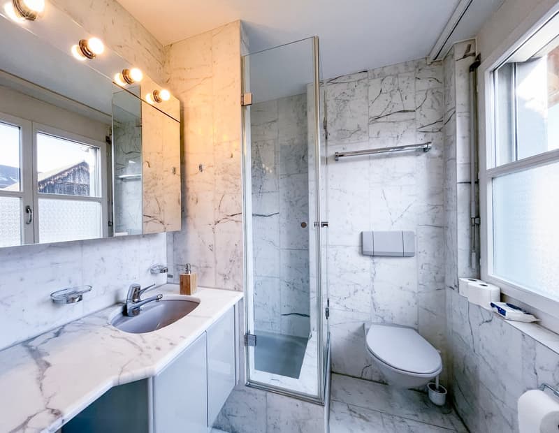 white marble tiles, glass shower enclosure, white sink, white toilet, white cabinets, mirror, toilet paper holder, soap dispenser, toilet paper