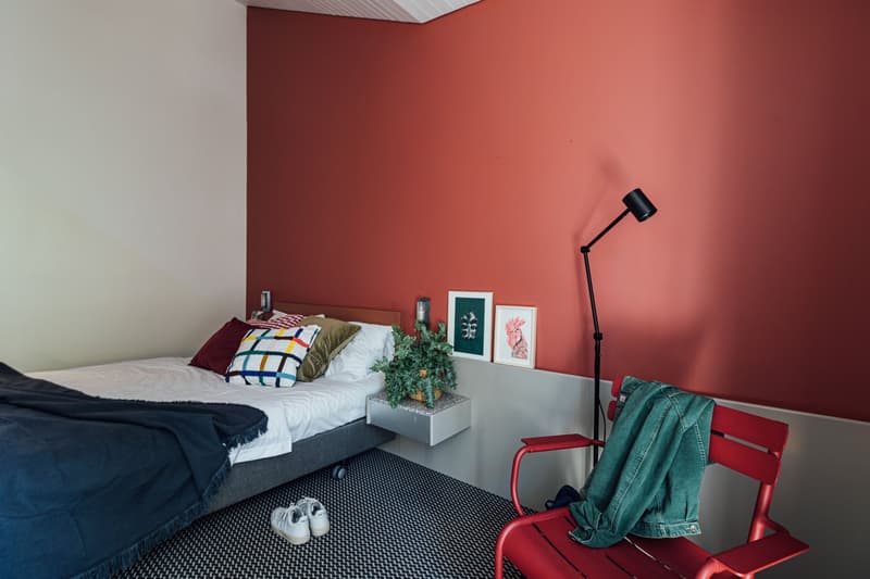 A small bedroom with a bed, a nightstand, a chair, a potted plant, and a wall lamp