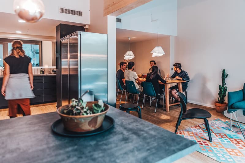 Open-plan living room and kitchen area with a dining table, modern appliances, and a glass table in the center. Wooden floors, white walls, and hanging lamps.