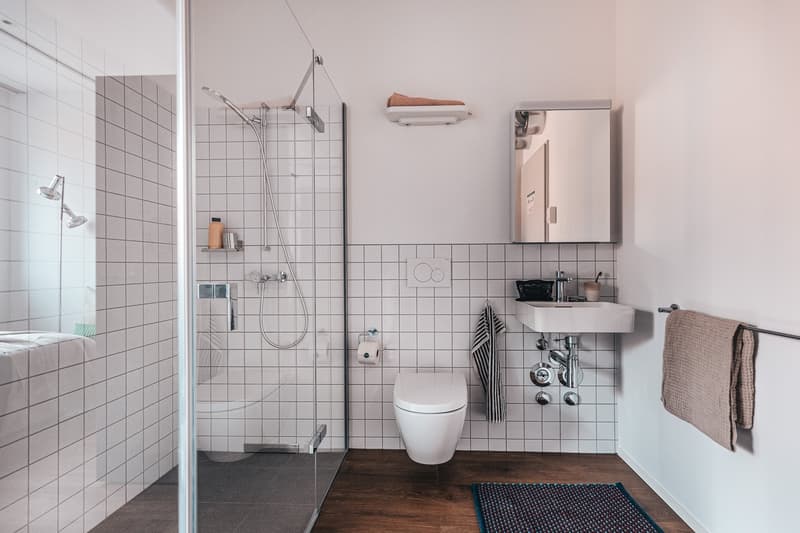 A modern, minimalist bathroom with white tiles, glass shower enclosure, white fixtures, towel rack, and a white toilet.