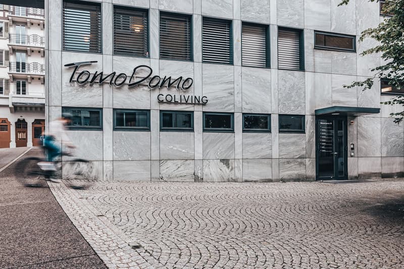 Contemporary building with modern design, large sign 'TomoDomo Coliving', windows with blinds, paved entrance, metallic door.