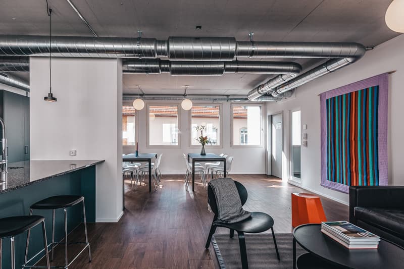 Open plan living and dining area with exposed ceiling ducts, a kitchen island, modern furniture, and a table with chairs.