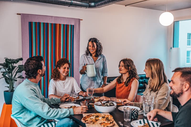 dining table, chairs, pizza, wine glasses, woman pouring water, people sitting