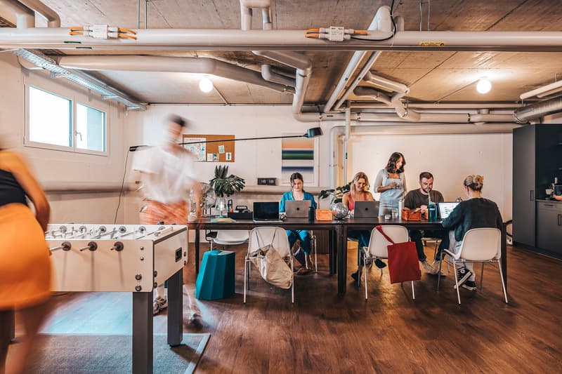 Room with desk, laptops, office chairs, people, foosball table, potted plants, window, radiators, white walls, wooden floor, storage unit, bulletin board.