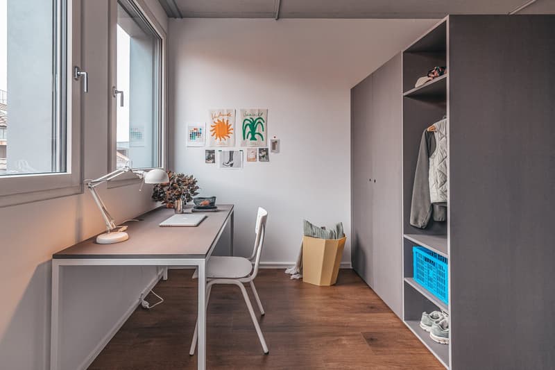 Small room, desk, white chair, wardrobe, window with view, table lamp, plant, storage box, pile of clothes