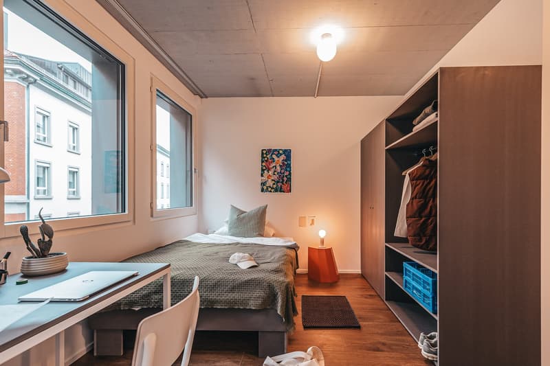 A compact room with a single bed, desk, chair, and a window with view of the outside. The room features wooden flooring and white walls. The desk has a laptop, and the bed is made with a blanket and a pillow. A nightstand with a lamp is beside the bed. A
