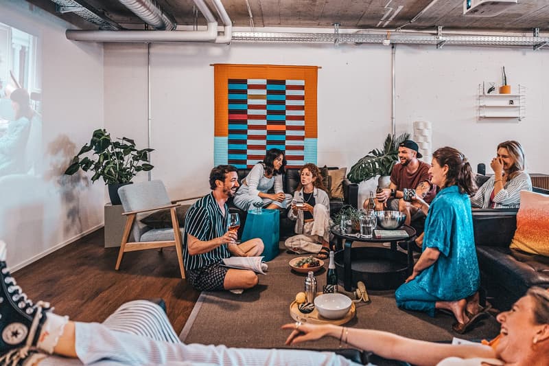 Common room: multiple people sitting on chairs, plants, coffee table, projector, table with wine glasses, bottles and other drinks.