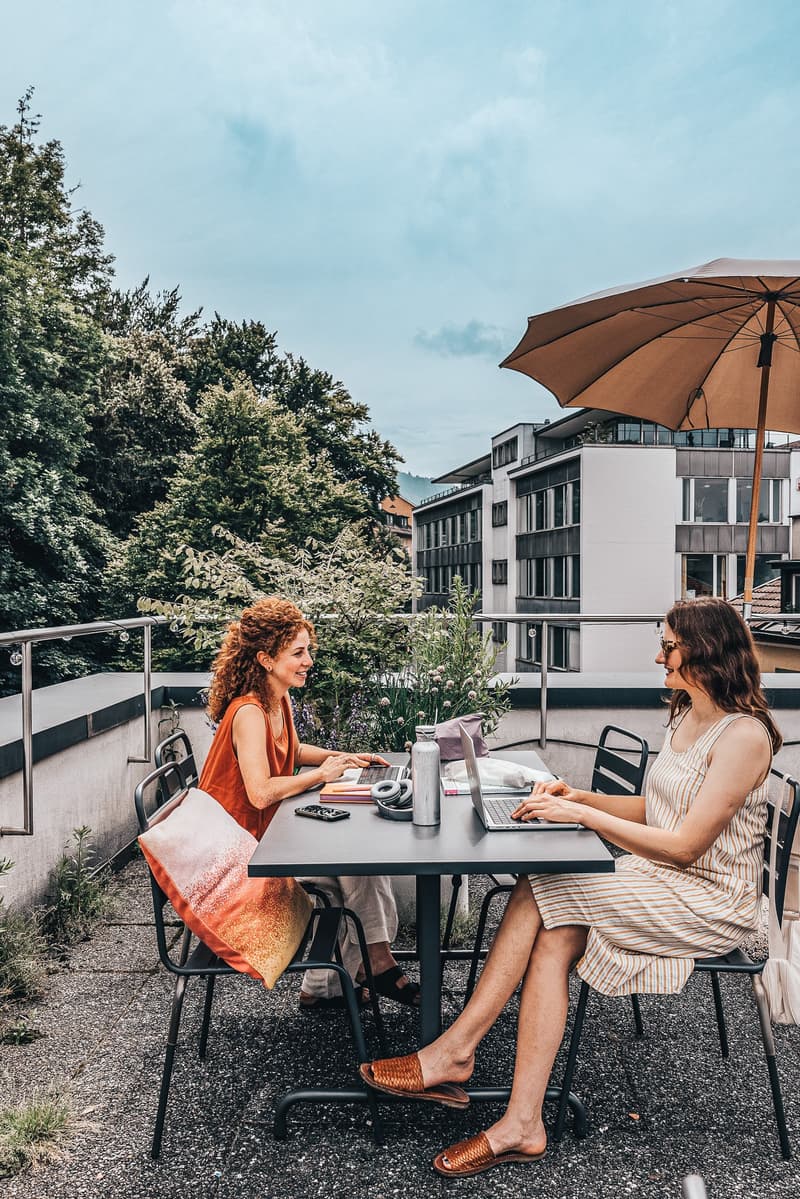 Outdoor terrace, metal table, black metal chairs, women working on laptops, open area with railings, trees and plants in the background