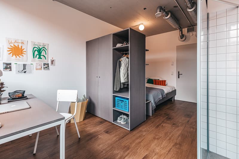 Bedroom with desk, gray wardrobe, bed, white chair, basket, laptop, plant, posters