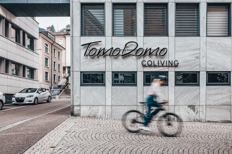 Stone facade building with the sign 'TomoDono Coliving', several floors with windows, a street with parked cars, a person riding a bicycle