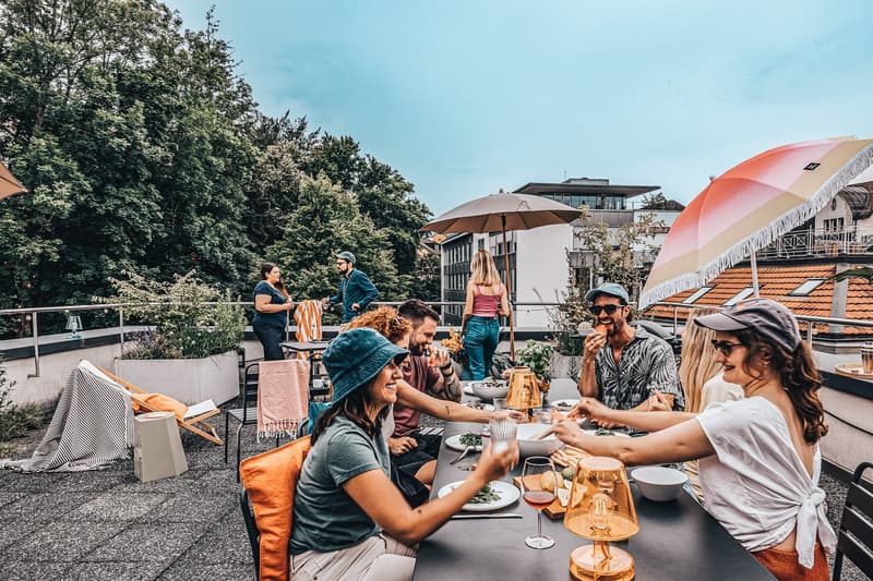 outdoor terrace with a long table, people sitting with open umbrellas, some standing near railings, trees and buildings in the background, a potted plant in the corner