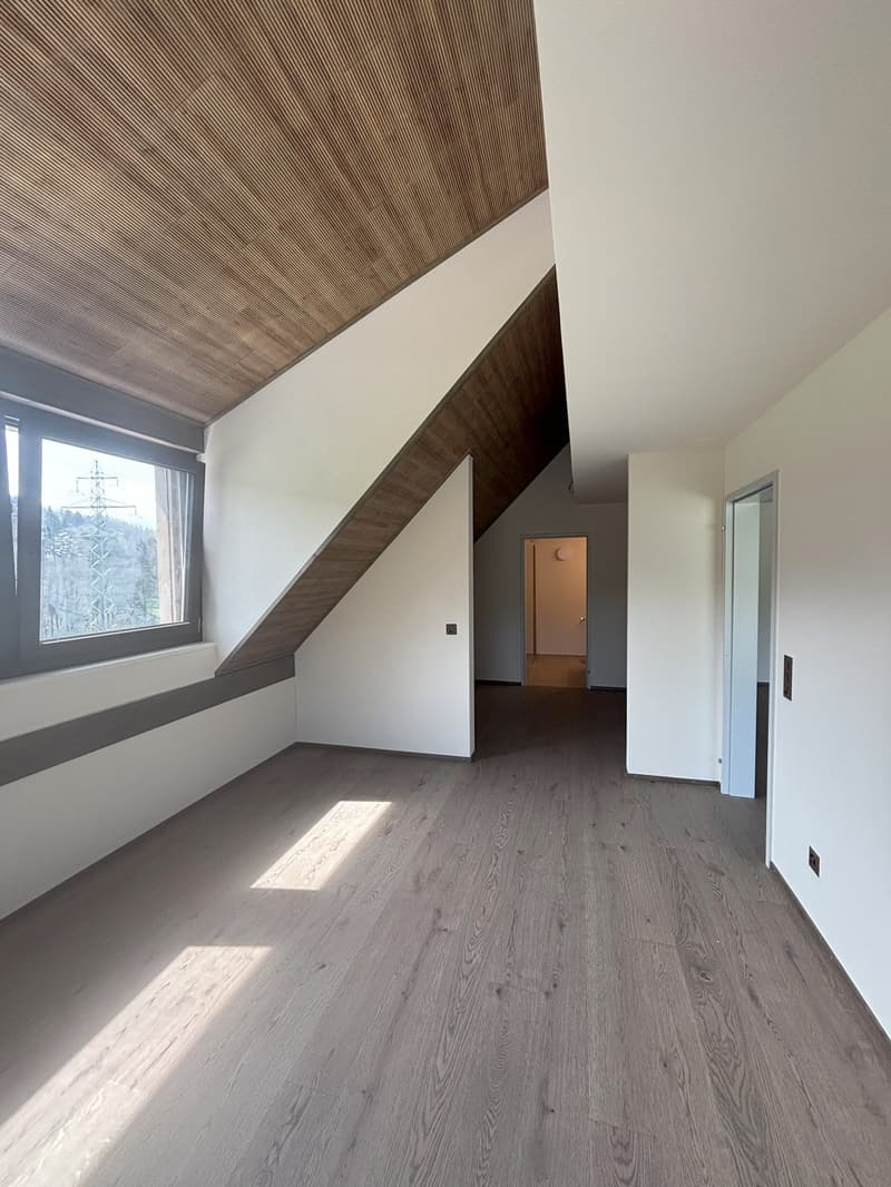 The image shows a room with a slanted wooden ceiling, white walls, and a large window. The floor is made of grey hardwood planks. The room has a minimalist and modern design aesthetic.