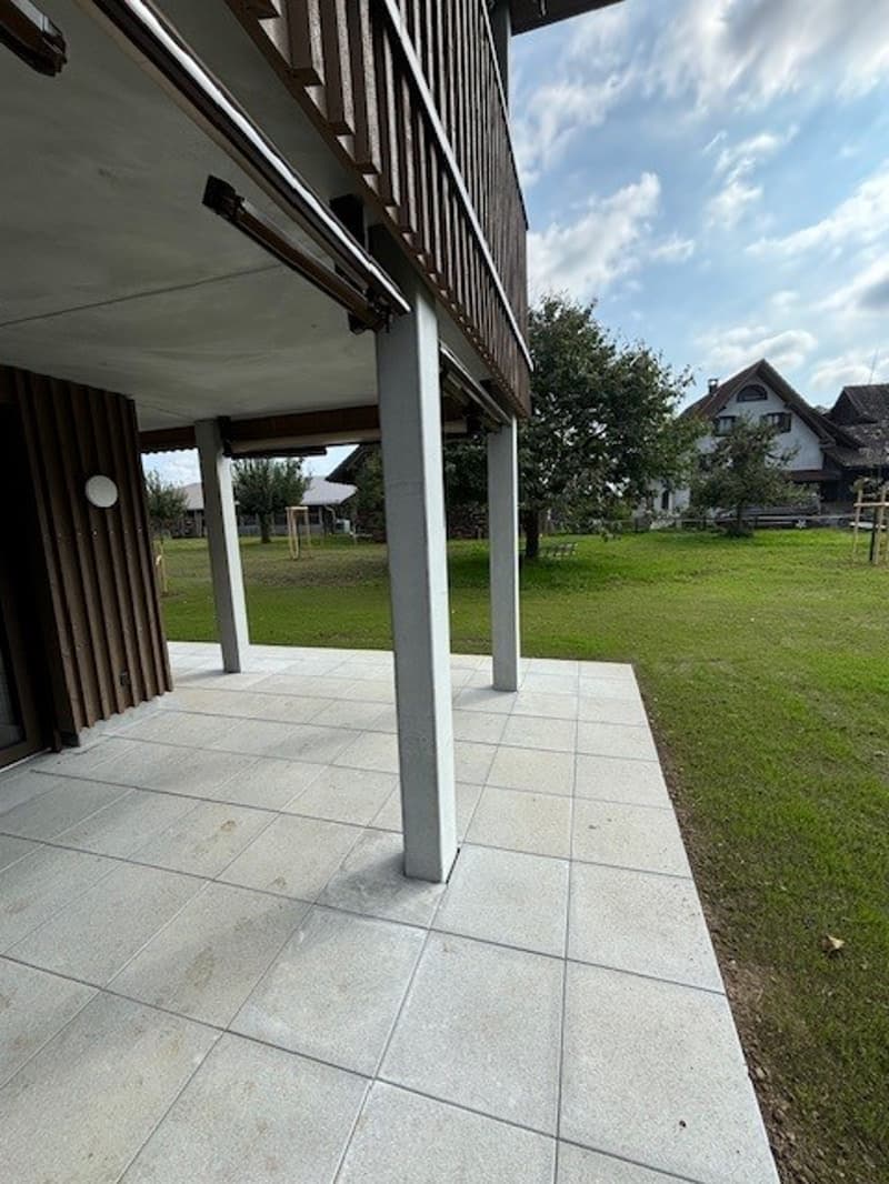 Raised ground floor, tiled floor, wooden wall, balcony, outdoor lighting, grassy field, trees, houses in the distance