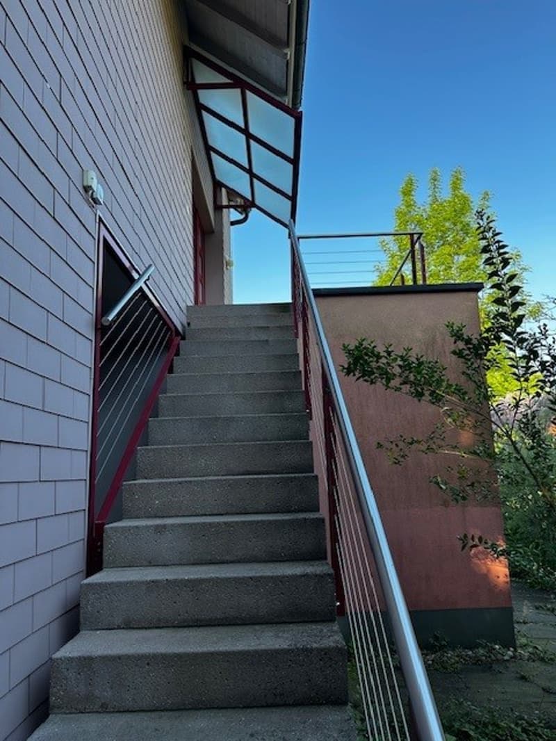 Exterior view of a multi-storey building, stairs leading to an upper floor, red handrails, tiled wall, and trees in the background