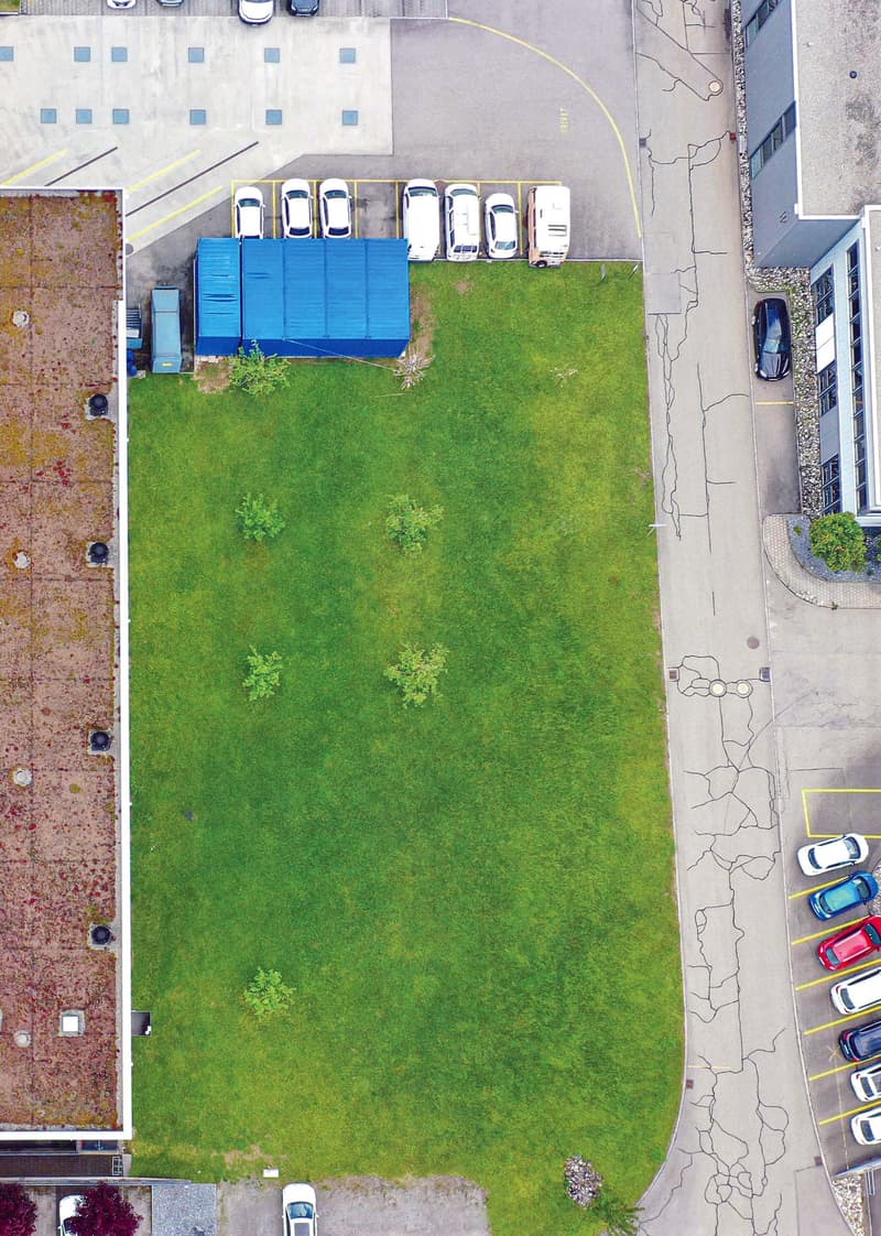 The image shows an aerial view of a parking lot with several parked cars. There is a grassy area in the center of the parking lot, with some small plants or shrubs visible. The parking lot is surrounded by buildings and other structures.