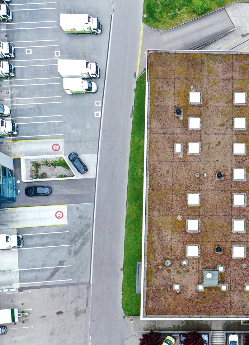 The image shows an aerial view of a parking lot. The lot is paved and has several marked parking spaces. There are some vehicles parked in the lot. The surrounding area includes some greenery, such as grass and trees.