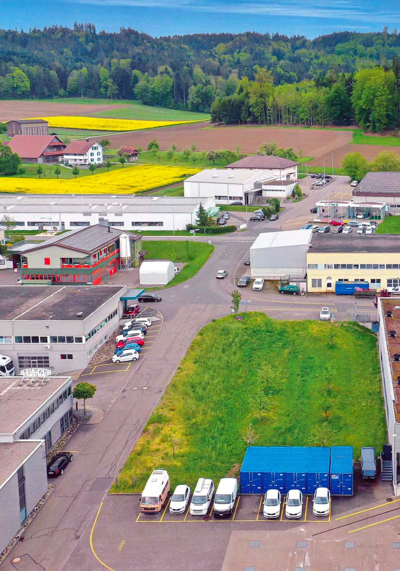 The image shows an aerial view of an industrial or commercial area. There are several buildings, including warehouses and office buildings, surrounded by paved parking lots and green spaces. The landscape includes fields with yellow flowers, forests, and