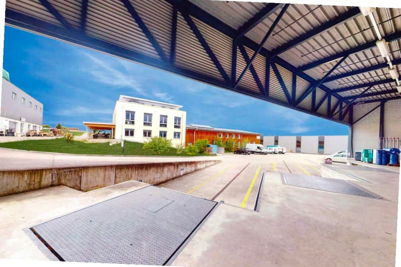 Large commercial building with a warehouse-like structure, metal roof, and multiple floors. There is a paved parking area in the foreground with yellow lines marking the spaces.