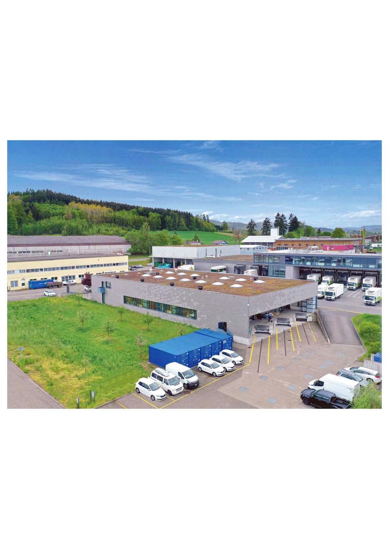 Large commercial building with a green roof, surrounded by a parking lot with several vehicles. The building appears to be a warehouse or distribution center, with loading docks and trucks visible. The surrounding landscape includes forested hills in the