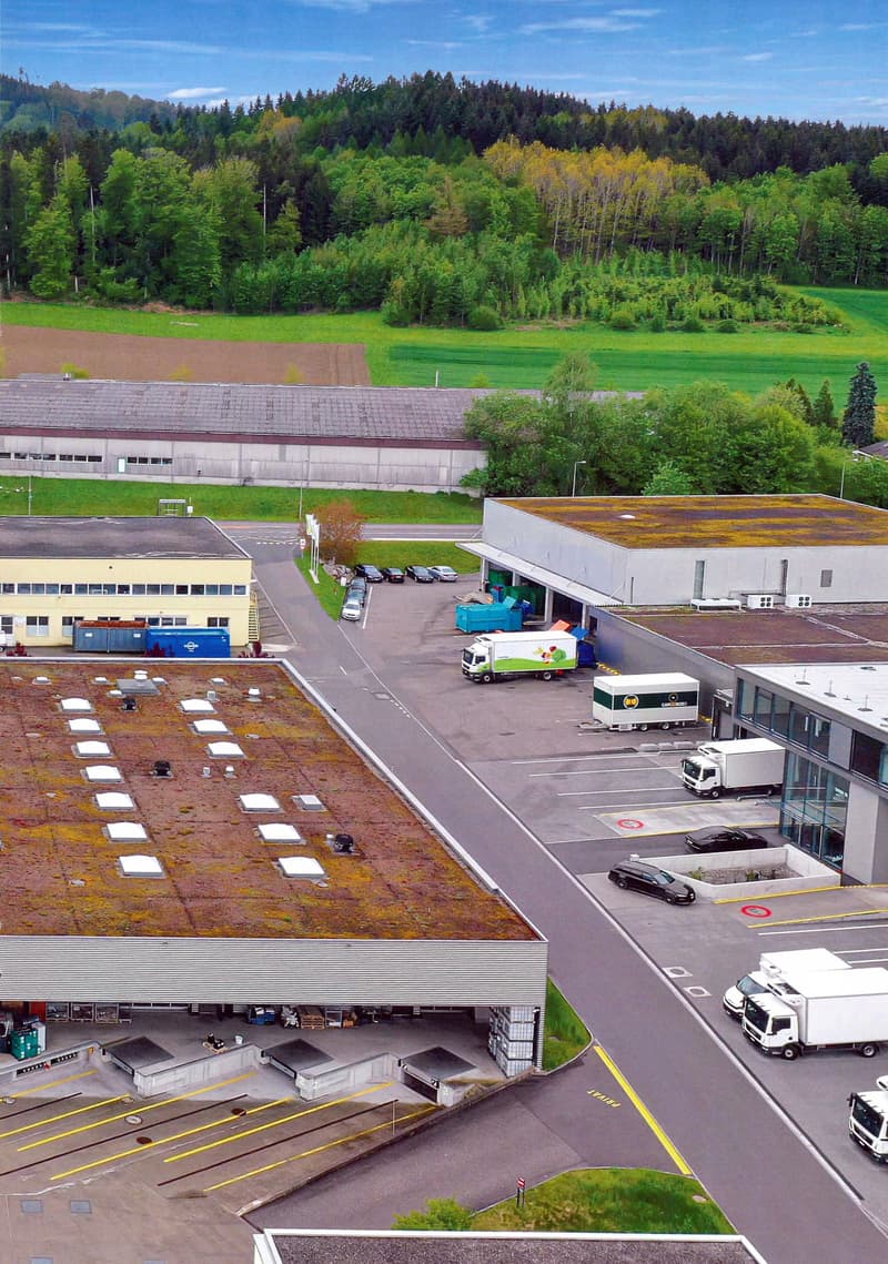 The image shows a large industrial or commercial building complex with a parking lot and loading docks. The building has a green roof and is surrounded by a forested area with trees in various shades of green and yellow, indicating a scenic, rural setting