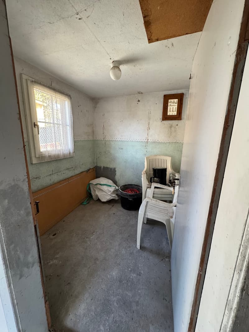 Storage room, white walls, ceiling lamp, wooden radiator, white window with bars and curtains, dirty floor, storage box and plastic bag on the floor