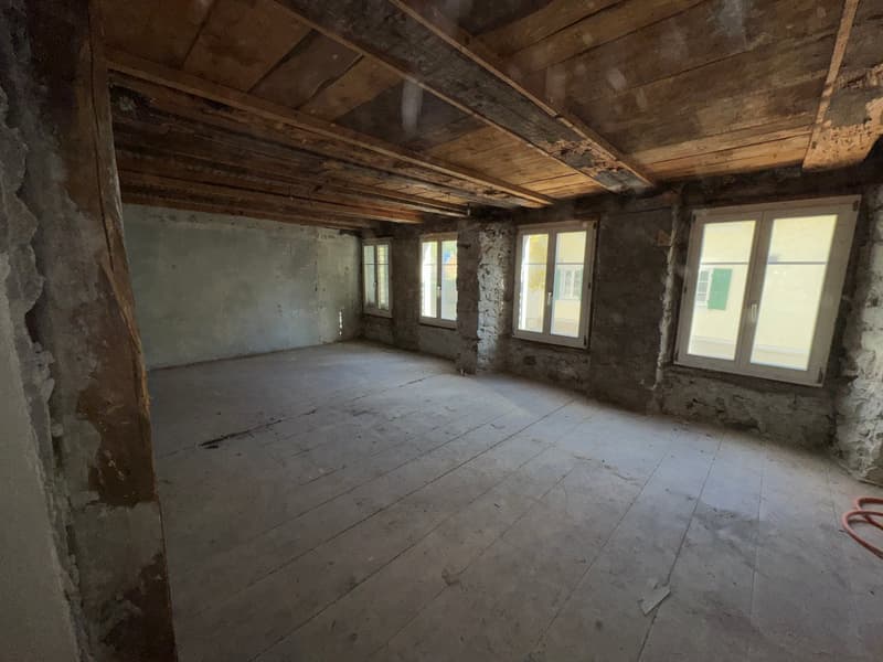 A large empty room with wooden floor, visible wood beams on the ceiling, stone walls, windows with shutters